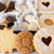 An assortment of cookies arranged in a box: round and star-shaped cookies dusted with powdered sugar, chocolate-covered cookies, and cookies with heart-shaped jam centers, alongside small rolled cookies in white paper cups.