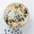 A top-down view of a round blueberry crumb cake topped with powdered sugar, with scattered blueberries on a white surface below.