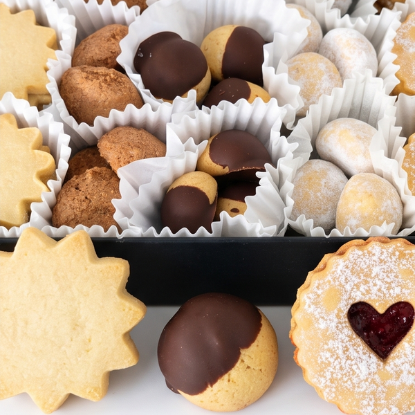 An assortment of Viennese-style cookies arranged in white paper cups inside a black box, with a few cookies placed in front on a white surface.