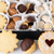 An assortment of Viennese-style cookies arranged in white paper cups inside a black box, with a few cookies placed in front on a white surface.