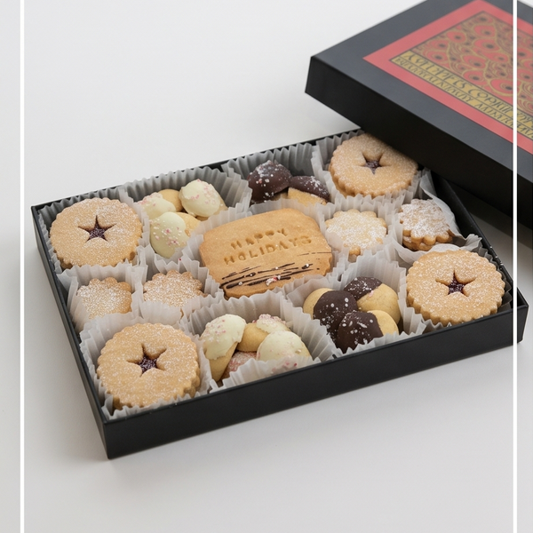 An open black gift box filled with assorted holiday cookies, including star-shaped linzer cookies, chocolate-dipped pieces, and a central biscuit stamped with 'Happy HOLIDAYS'. The matching lid rests beside the box on a clean white background.