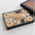 An open black gift box filled with assorted holiday cookies, including star-shaped linzer cookies, chocolate-dipped pieces, and a central biscuit stamped with 'Happy HOLIDAYS'. The matching lid rests beside the box on a clean white background.