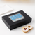 A black rectangular gift box shown on a white surface with two powdered-sugar-dusted Linzer cookies in front.