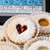 Heart-shaped Linzer cookies dusted with powdered sugar, with one cookie showing a red jam center and another with a yellow center, arranged in white paper cups with a Vienna Cookie Company sign in the background.